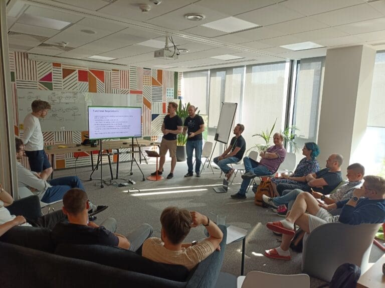 Blog visual: A group of professionals in a modern office space is engaged in a presentation. Two presenters stand near a screen displaying "Functional Requirements," while attendees sit in a semi-circle, attentively listening. The setting features large windows allowing natural light, a whiteboard with diagrams, and a colorful geometric-patterned wall. Some participants are taking notes, while others lean back, reflecting on the discussion. The atmosphere suggests a collaborative workshop or brainstorming session.